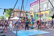 Street performers execute an acrobatic act before a large crowd at an outdoor festival, with murals and tents in the background.