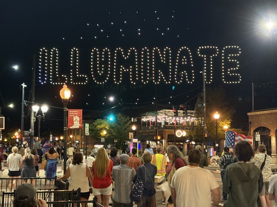 A crowd watches as drones form the word “ILLUMINATE” in glowing letters above a city street at night.