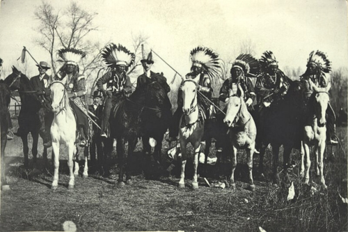a group of Ute Indians on horseback in full regalia with head dresses