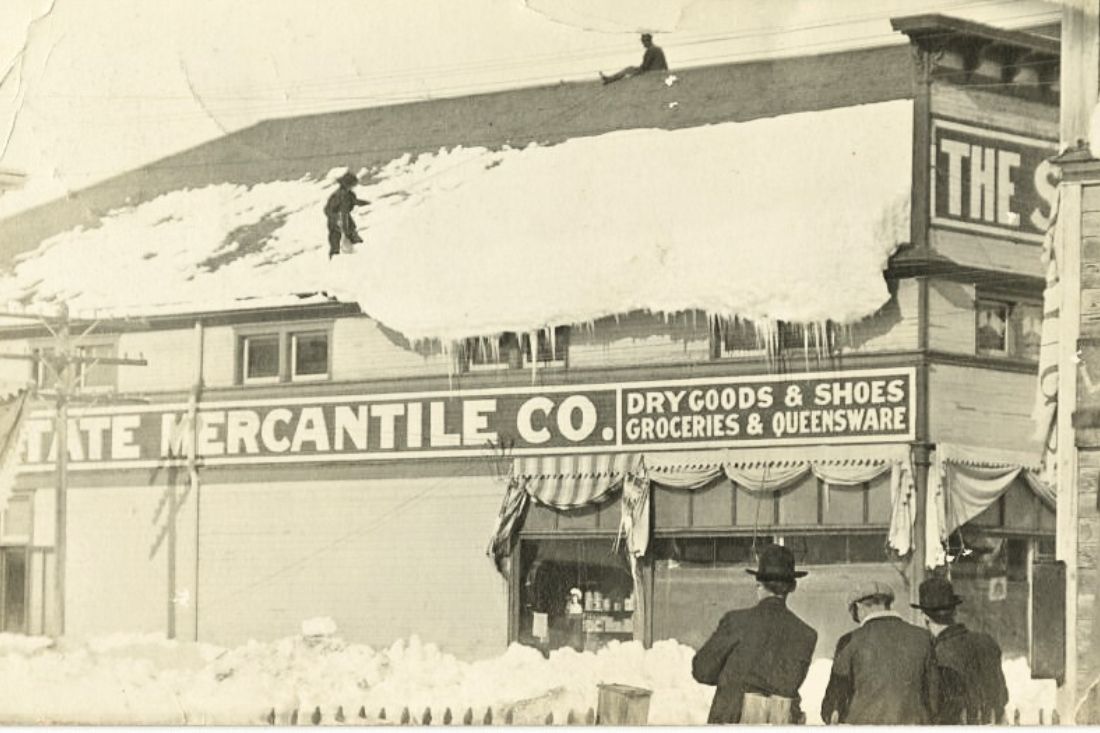 State Mercantile building with a roof covered in heavy snow that is falling off, two men are on roof. 