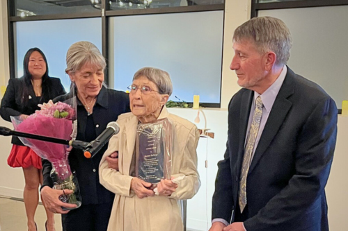 a woman talks into a microphone holding flowers, with her adult children on either side of her