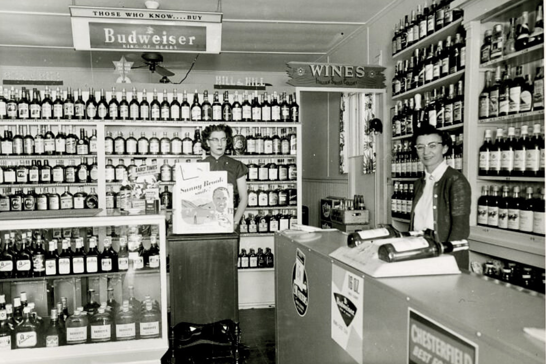 Two women stand behind the counter at a liquor store. Many bottles line the walls behind them.