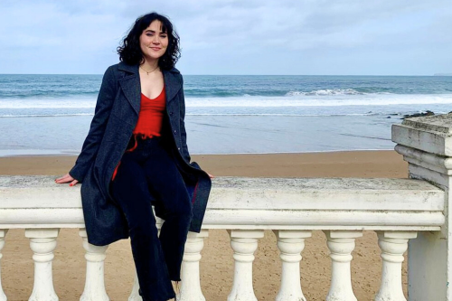 a woman with dark hair sits on a railing in front of a blue sea