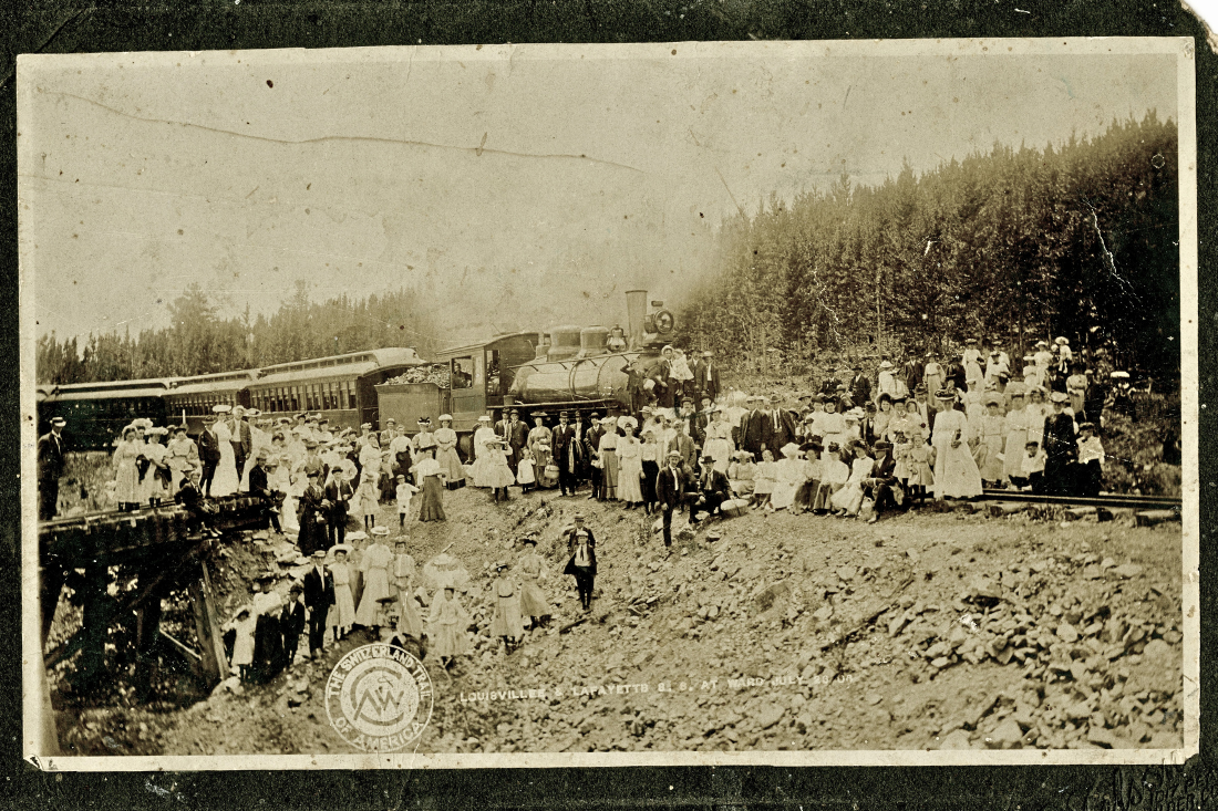 a large group of men and women stand in front of a steam engine train in the mountains.