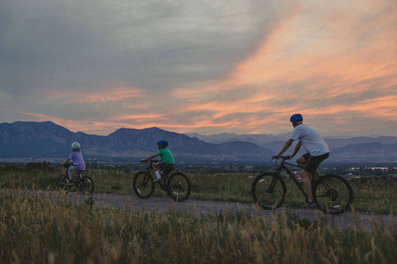 an adult and two kids riding along a trail in Louisville with a beautiful sunset behind the flatirons in the distance