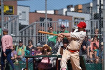 Man swinging baseball bat in vintage baseball uniform. There is a crowd of spectators watching from the stands in the backgroud.