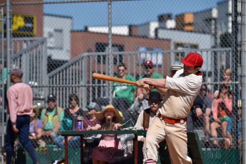 Man swinging a baseball bat in a vintage baseball uniform. There is a crowd of spectators watching in the stands behind him.