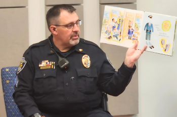 A police officer in uniform sits and reads a children's book aloud, holding the book open to show the illustrations.
