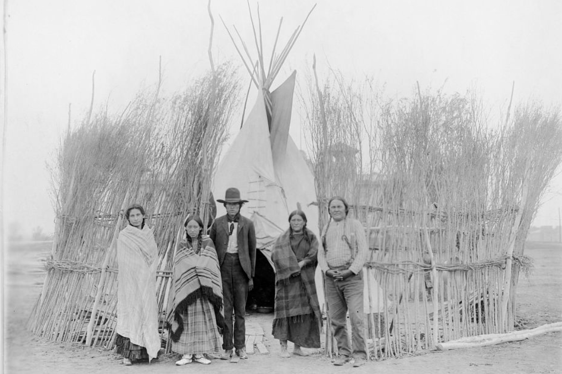 Black and white photo of five Arapaho standing in front of tipi surrounded by a brush fence. 