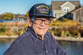 An older man with glasses and a Veteran hat stands by a river, enjoying the serene natural surroundings.