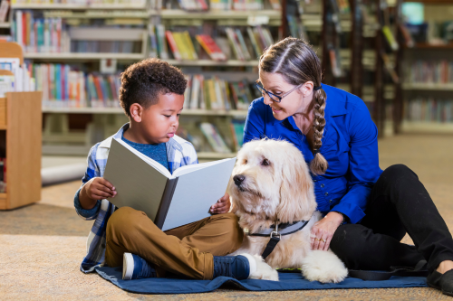 A young boy reads a book to a woman and a therapy dog while sitting on the floor in a library.
