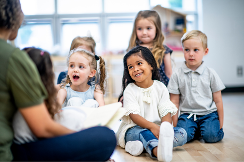 group of young children sitting on the floor listening to a teacher read a book