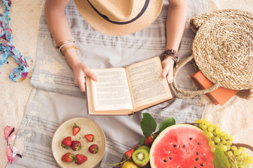 looking from above of person reading book on the beach with beach towel, fruit, and sunglasses around