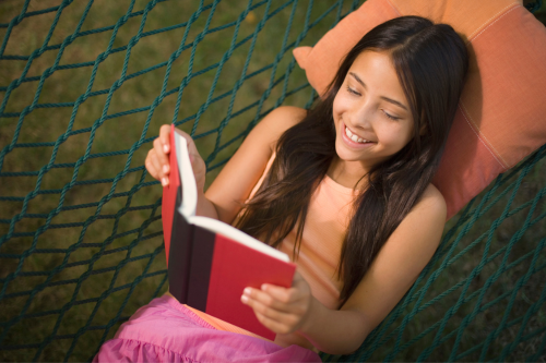 teen girl laying in a hammock outside reading a book