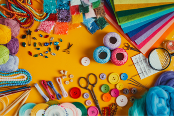 Colorful craft supplies arranged on a bright yellow background: yarn, beads, buttons, scissors, thread, fabric swatches, and a tape measure.