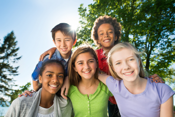 Five diverse, smiling teenagers pose closely outdoors beneath leafy trees, arms around one another, conveying friendship, joy, and unity.