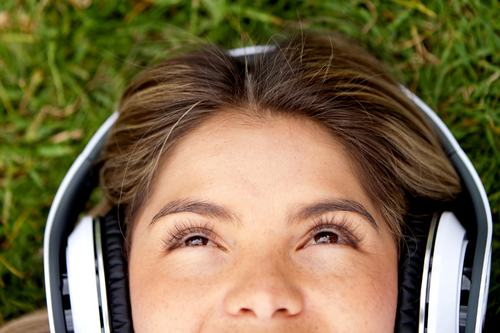 Close-up of a smiling teen lying on green grass, wearing large white headphones and gazing upward in a relaxed, peaceful mood.