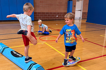 Kid standing on a foam crayon, and a kid standing on a balance ball for a sports action class