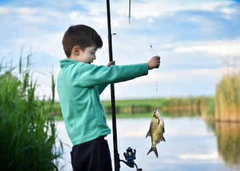 A young boy holds a fishing pole with a fish in front of body of water