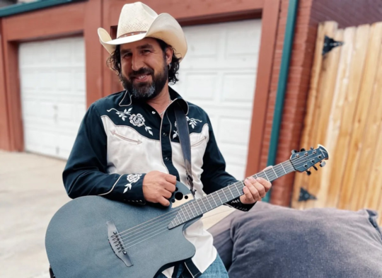 Musician Aaron Rose poses in a cowboy hat with a blue guitar