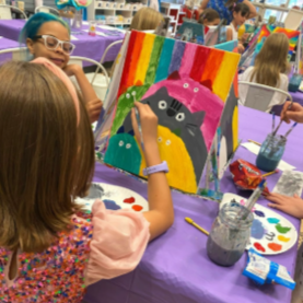 girl painting a pic of a bunch of cats on a rainbow background on a purple table