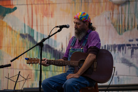 bearded man with a rainbow bandana, purple shirt and blue overalls plays guitar on stage at open mic night