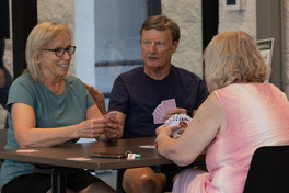 3 seniors sit at a table at the SCC playing euchre: a woman in a teal shirt, a man in a navy shirt, and a woman in a pink dress