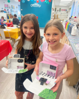 2 girls in art classroom holding dioramas of small businesses made from cardboard boxes and craft supplies