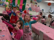 group of kids in an art classroom with pink tablecloths holding slime and stretching it for the camera