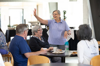 a woman in a purple shirt standing at a table that has 3 seniors sitting at it waves to someone else off camera