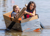 Two girls paddle in a cardboard boat on a body of water