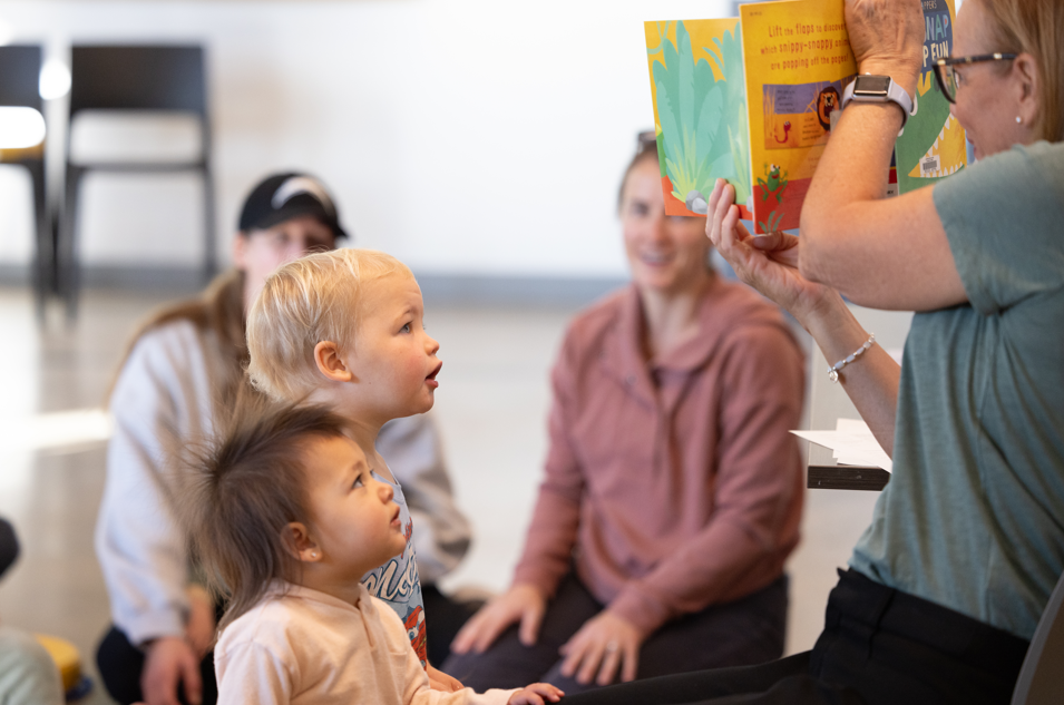 2 children staring up at the SCC facilitator reading to them at family storytime while their parents smile in the background