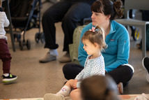 a woman in a blue jacket sits on the floor with a child in her lap listening to a presentation at the SCC