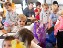toddler boy staring at camera amongst group of other toddlers while teacher passes out colorful ribbons to swirl