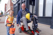 A young child in costume and a man in a motorized wheelchair trick-or-treat.