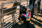 A man in a jacket and sunglasses and a child in a tie-dye jacket feed a goat