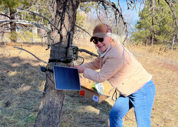 A woman in a tan jacket and hat smiles while adjusting a wildlife monitoring camera