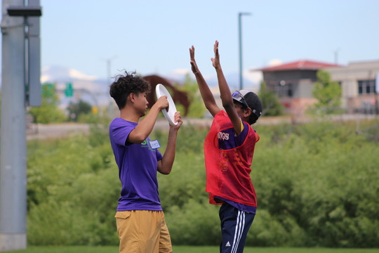 A kid in a purple shirt holds a white frisbee while another kid lifts their arms