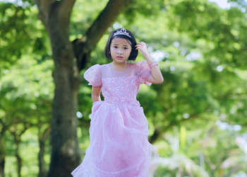 A young girl stands outside in a silver crown and a pink dress 