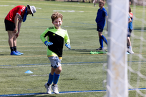 A young boy in a green and black shirt and blue gloves runs on a field during a soccer camp