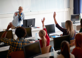 A teacher with short white hair and a blue shirt speaks to a class with students who are raising their hands