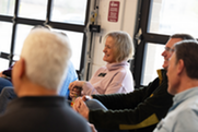 A woman with blonde hair and a purple turtleneck smiles in a group of people during First Fridays Coffee