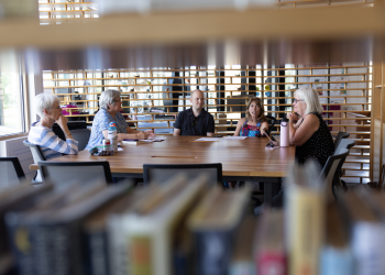 Five people discuss books around a table as seen through library bookshelves