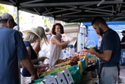 Person smiling while grabbing a produce bag and leaning over tables stocked with colorful fruits and vegetables during a market.
