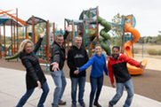 Five people pose with big smiles and outstretched arms in front of the new community park playground equipment