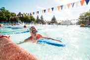 A kid swims in the pool with a blue kickboard while wearing goggles and a pink swim cap with kiwi fruit 