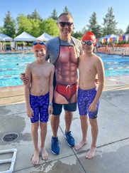 Two boys in blue swimsuits and orange swimcaps and a man wearing sunglasses and an apron with abs smile for a photo in front of a pool