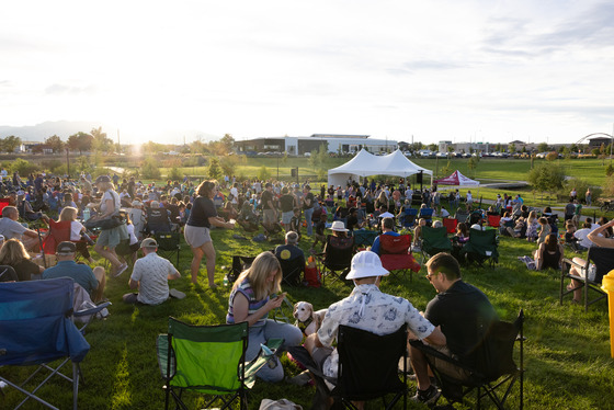 A crowd of people sit in chairs and blankets on the grass with a mountain backdrop while listening to live music during the Summer Concert Series