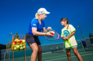An instructor in a blue shirt and hat places a tennis ball in front of a child's racket