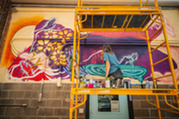 An artist stands on scaffolding while painting a mural on a city wall